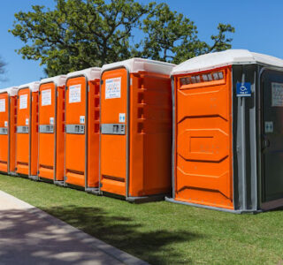 Row of orange portable toilets and one accessible toilet unit in a park on a sunny day.
