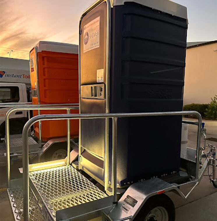 Portable shower unit on a trailer, illuminated at sunset, with another unit and a truck in the background.