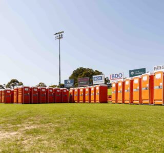 portable toilet odor control - Row of orange portable toilets lined up on a grassy field at a sports stadium under clear skies.
