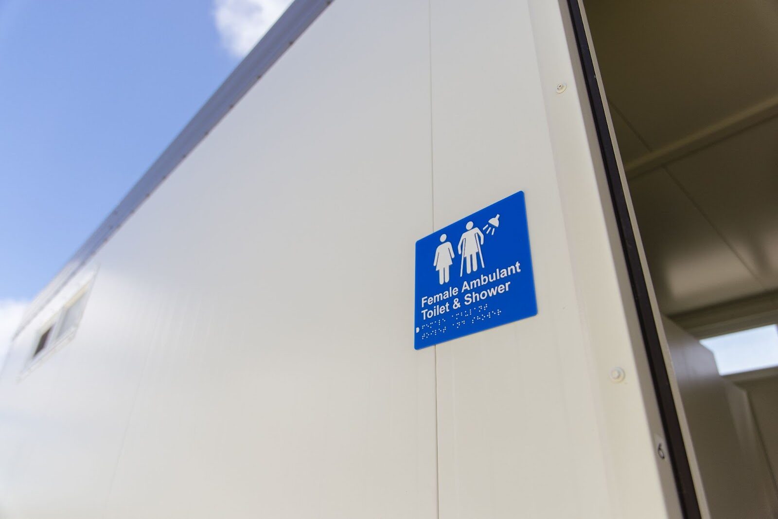 Close-up of a sign on a portable facility indicating Female Ambulant Toilet & Shower with accessibility symbols, mounted on a white exterior wall.