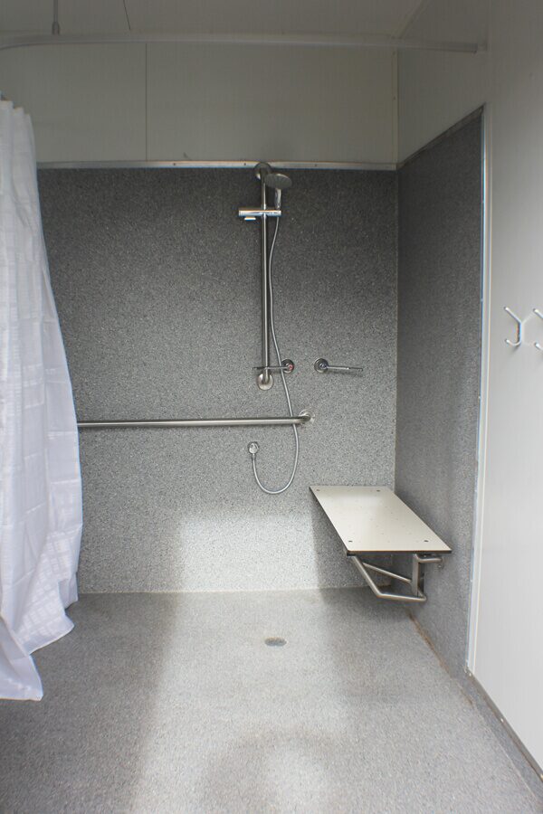 Vertical image of an interior shower stall with a metallic wall-mounted shower head, handle, and seat in a portable facility.