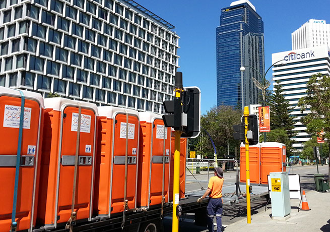 Row of orange portable restrooms being transported on a trailer in an urban area.