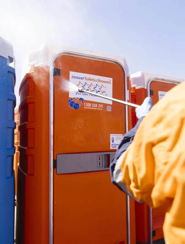 Worker cleaning a portable orange toilet from Instant Toilets & Showers with a power washer.