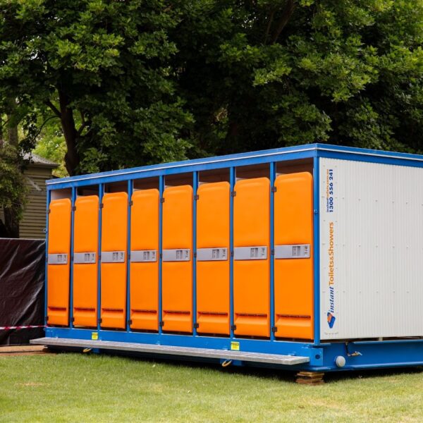 Portable restroom units with bright orange doors and blue frames placed outdoors on green grass, surrounded by trees.