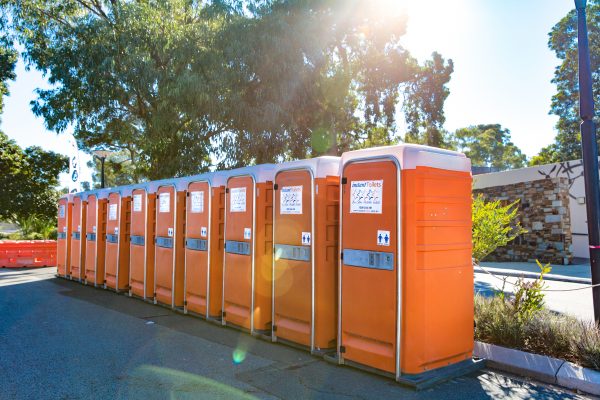 Row of orange Event portable toilet in an outdoor area with sunlight filtering through trees.