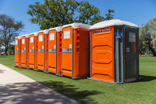 Row of orange portable toilet for weddings, including an accessible unit, set up on a grassy area.