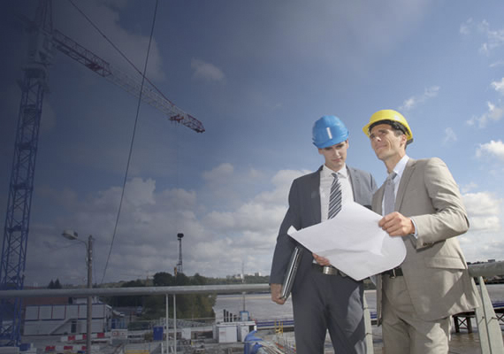 Two engineers in hard hats reviewing blueprints at a construction site with cranes in the background.