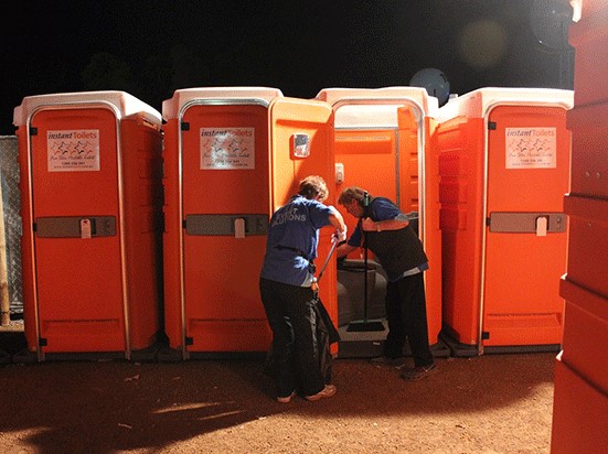 Two workers cleaning an orange portable toilet at night for portable toilet services with additional units in the background.