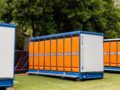 Portable restroom units with bright orange doors and blue frames placed outdoors on green grass, surrounded by trees.