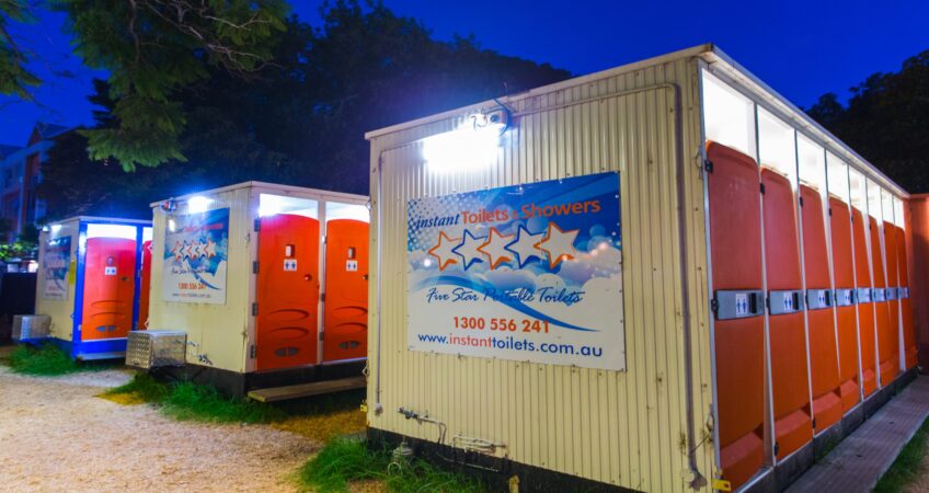 Portable toilets and showers setup at an outdoor event during the evening, illuminated by overhead lights.