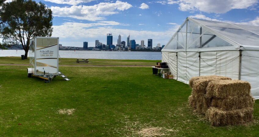 Outdoor event setup with a mobile toilet trailer, hay bales, and a marquee against a backdrop of a city skyline and waterfront.
