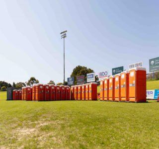 portable toilet odor control - Row of orange portable toilets lined up on a grassy field at a sports stadium under clear skies.