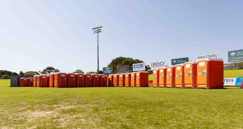 Row of orange portable toilets lined up on a grassy field at a sports stadium under clear skies.